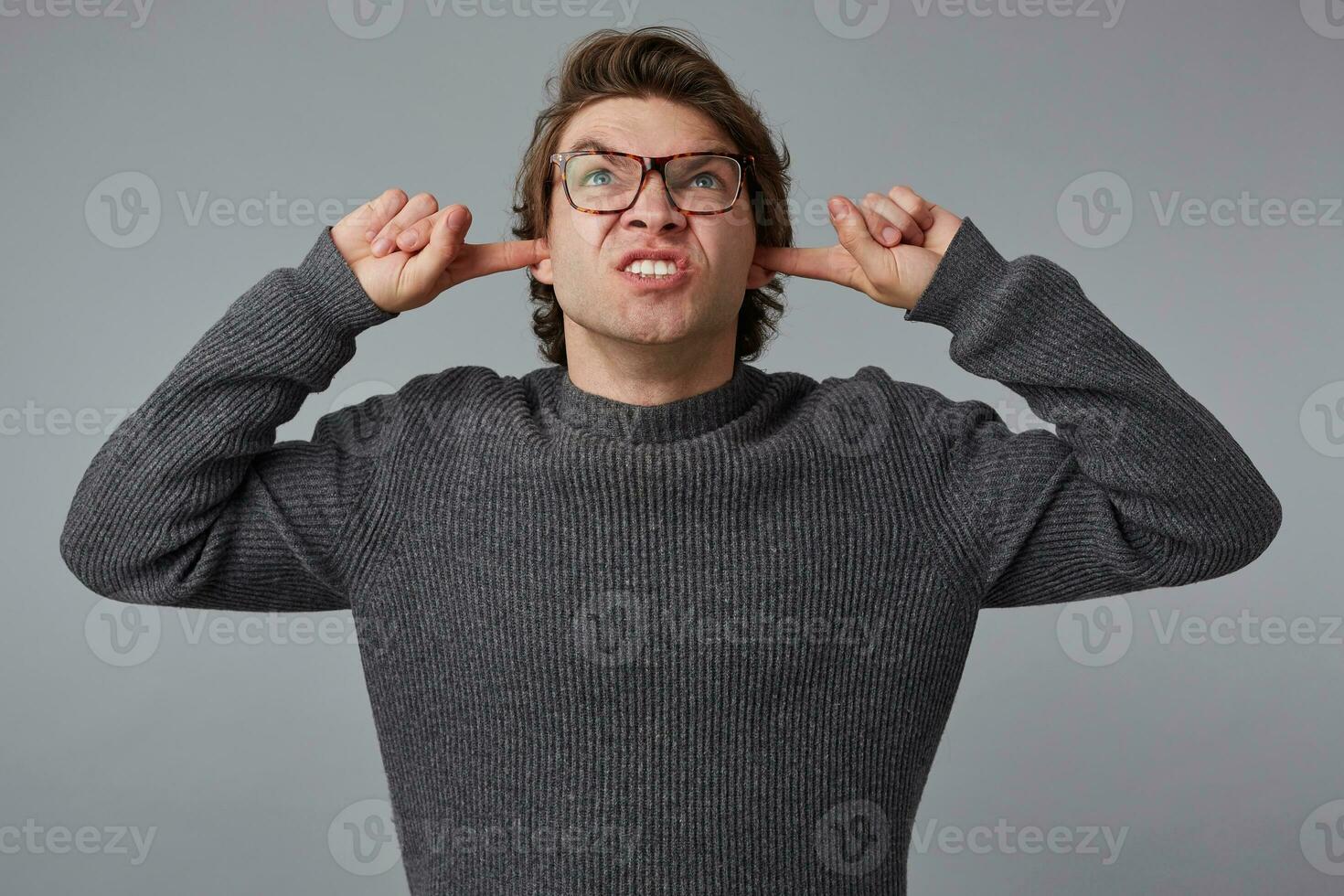 Portrait of young angry man with glasses wears in gray sweater, stands over gray background, disturbed by noise and covers ears with fingers. photo