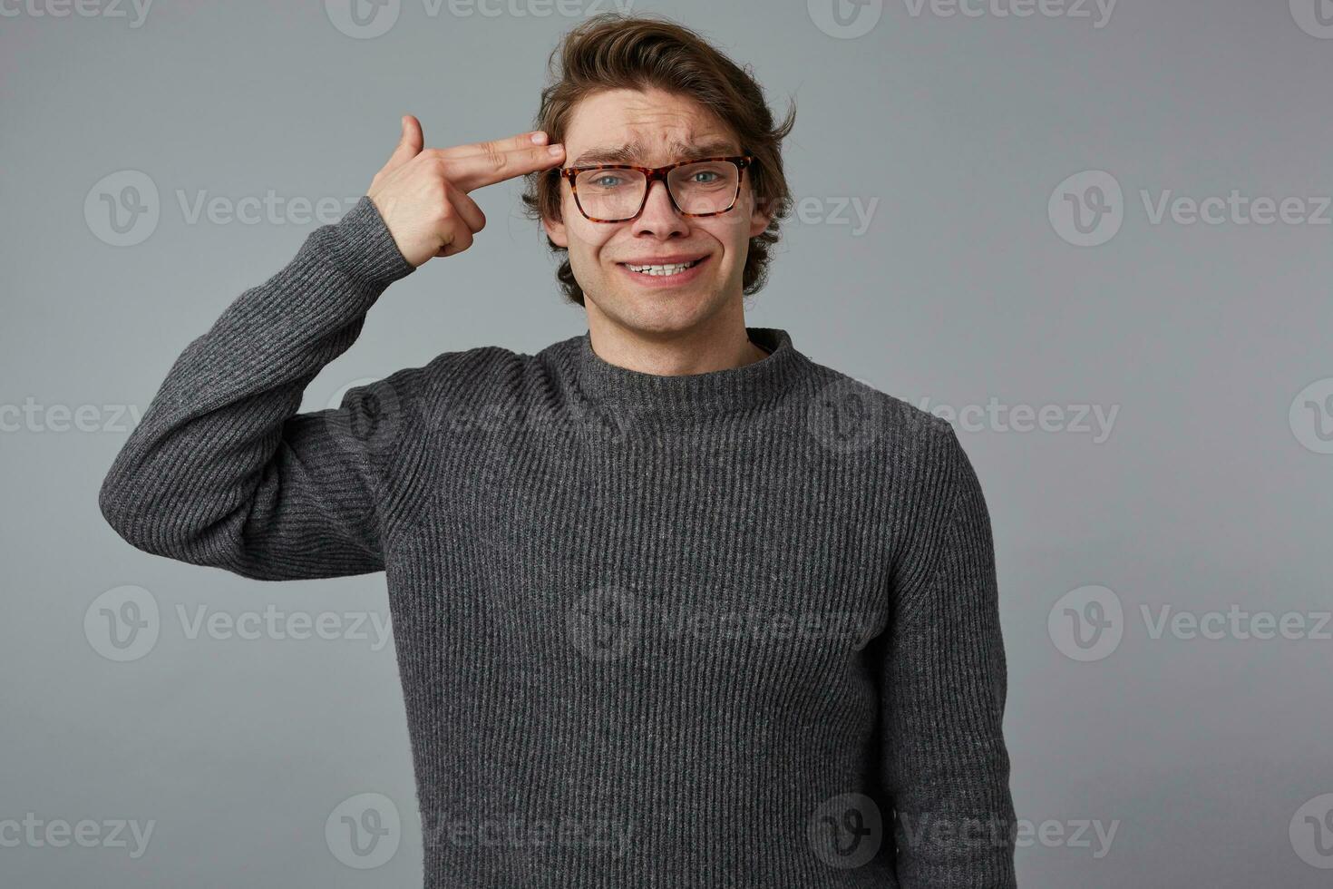 retrato de joven hombre con lentes usa en gris suéter, soportes terminado gris antecedentes gritos en templo con dedo, demuestra suicidio gesto. foto