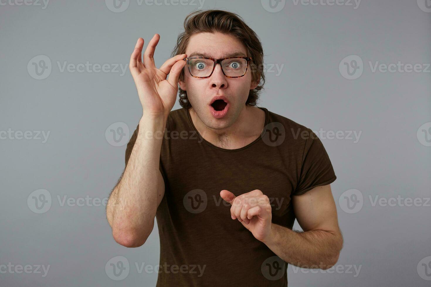 Young wondered with glasses wears in basic t-shirt, stands over gray background and broadly smiles, looks looks trough glasses with wide open mouth and eyes. photo