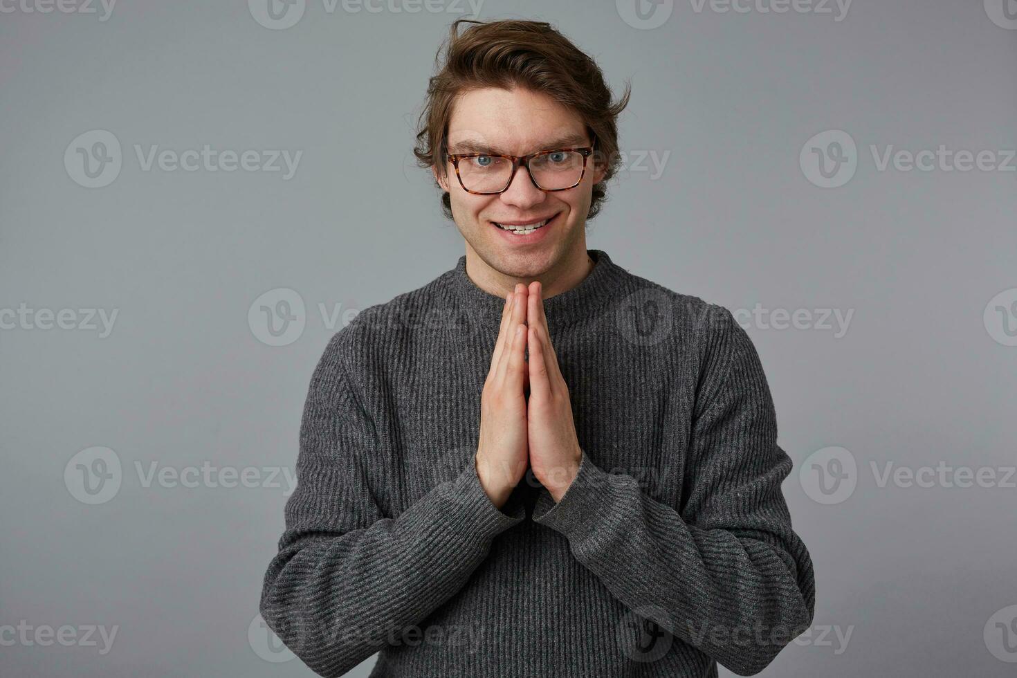 Portrait of young man with glasses wears in gray sweater, stands over gray background, it should be folded palm and insidiously looking into the camera. photo