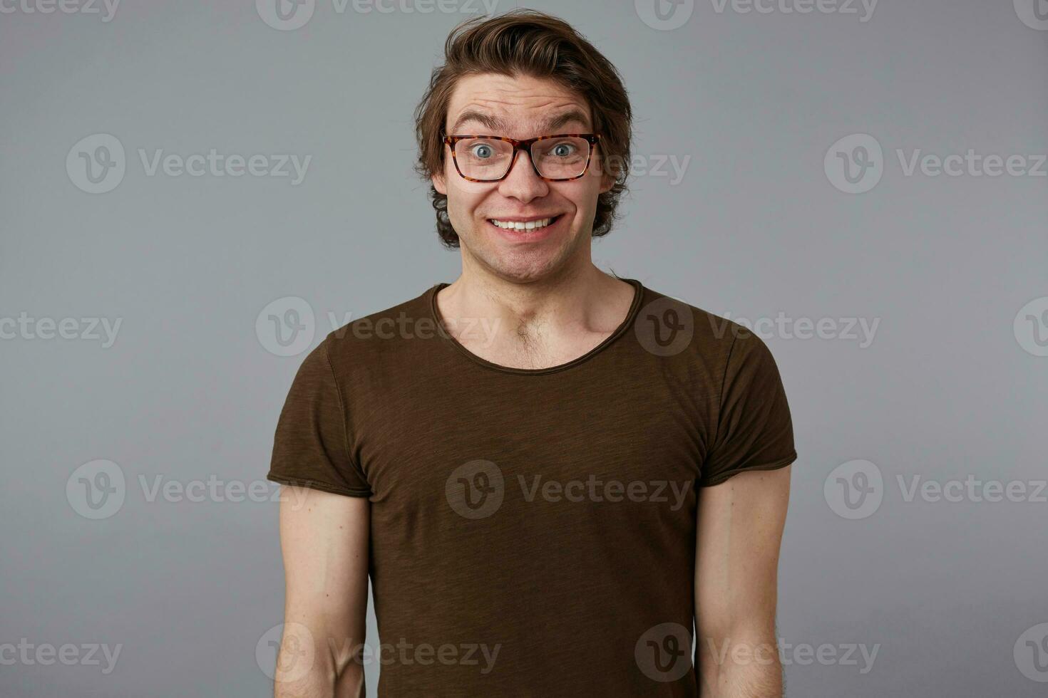 Photo of young surprised handsome man with glasses wears in basic t-shirts, stands over gray background and broadly smiles, looks happy and wondered.