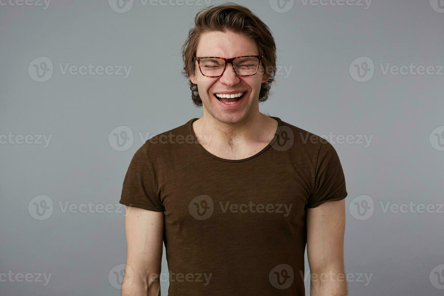 Photo of young loughting handsome man with glasses wears in basic t-shirt, stands over gray background and broadly smiles, looks happy and wondered.