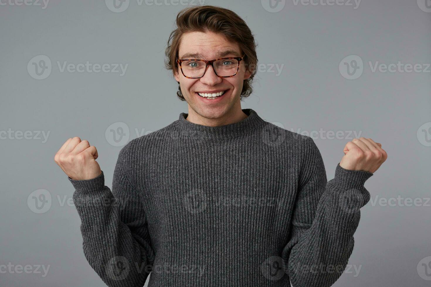 Young happy guy with glasses wears in gray sweater, stands over gray background. Broadly smiles and clenches fists, won the and feels great. photo
