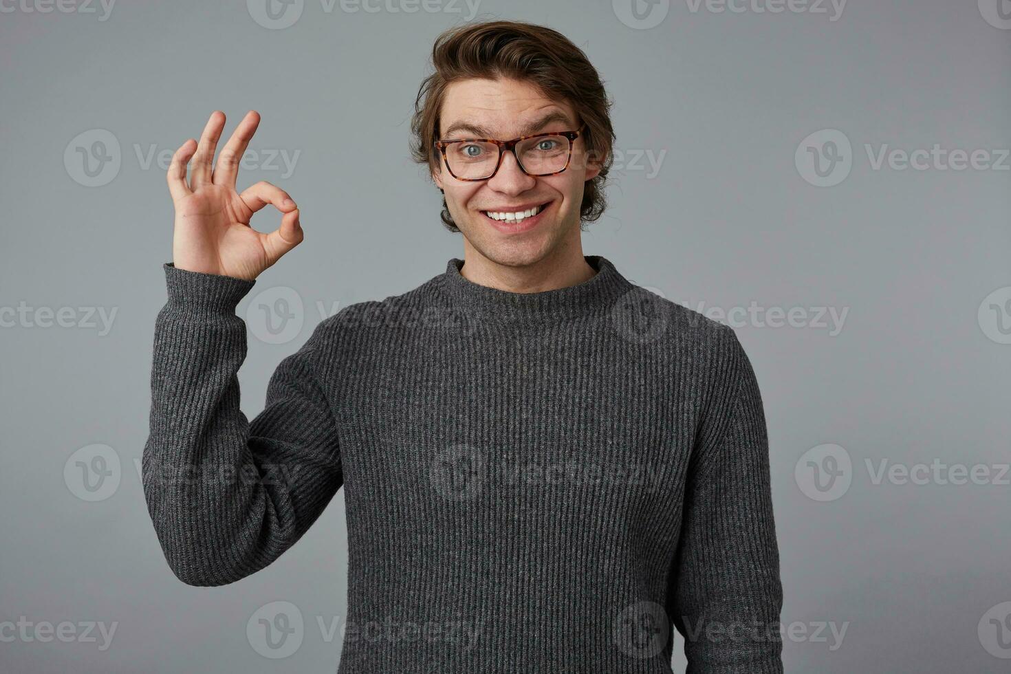 Photo of young cheerful handsome guy with glasses wears in gray sweater, stands over gray background and broadly smiles, looks happy and glad, shows okey gesture.