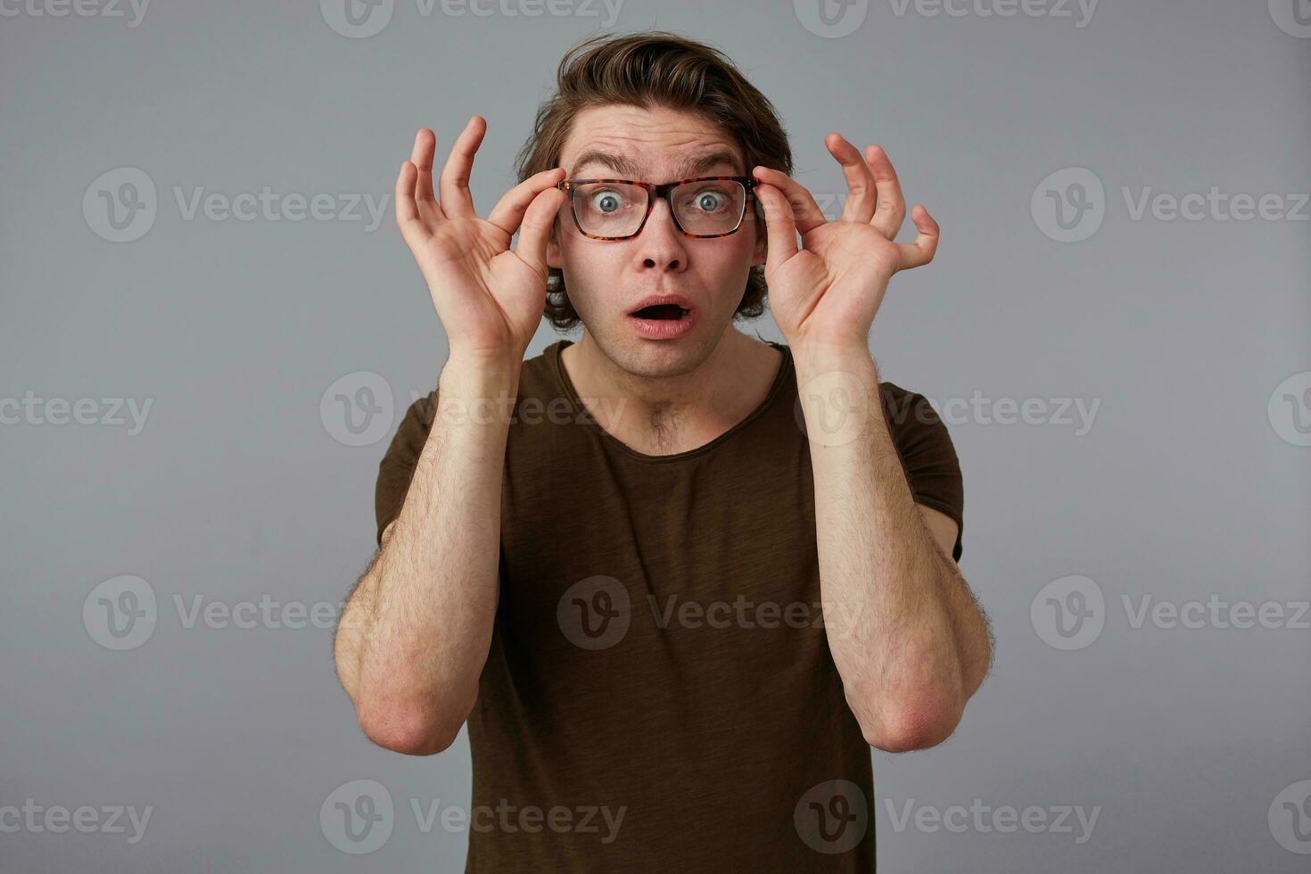 retrato de joven preguntado hermoso hombre con lentes usa en básico camiseta, soportes terminado gris antecedentes y en general sonrisas, mira mira canal lentes con amplio abierto boca. foto