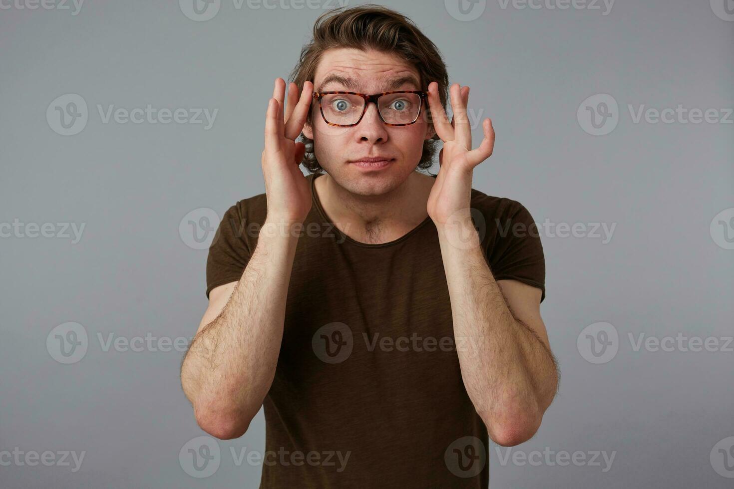 Portrait of young pensive man with glasses wears in basic t-shirt, stands over gray background and looks at the camera trough glasses. photo