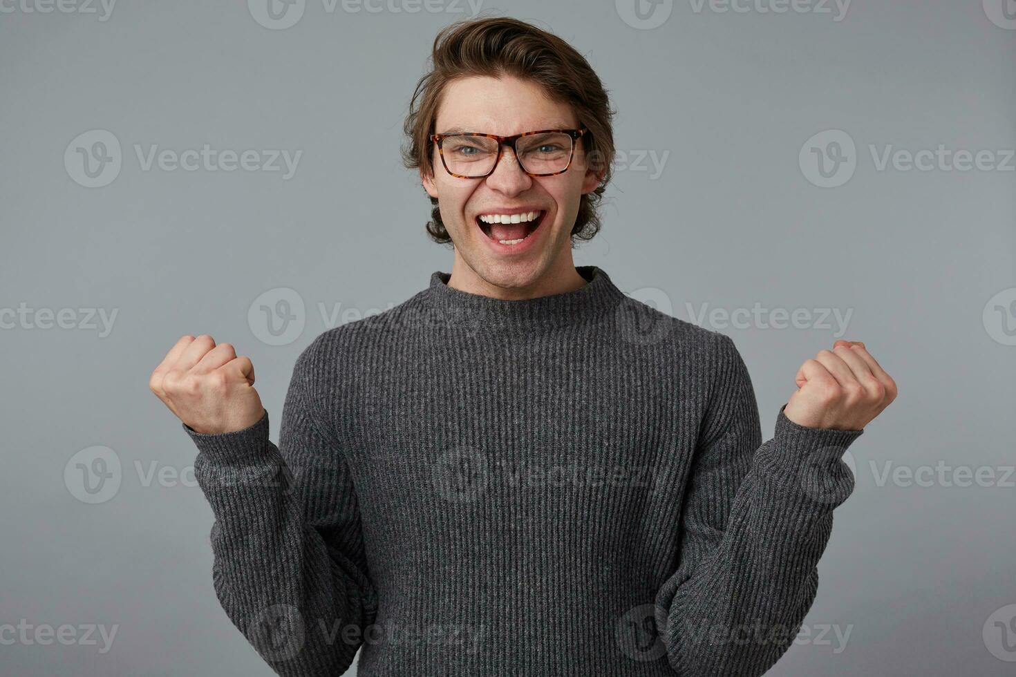 Portrait of young glad man with glasses wears in gray sweater, stands over gray background. Broadly smiles and clenches fists, won the car and feels happiness. photo