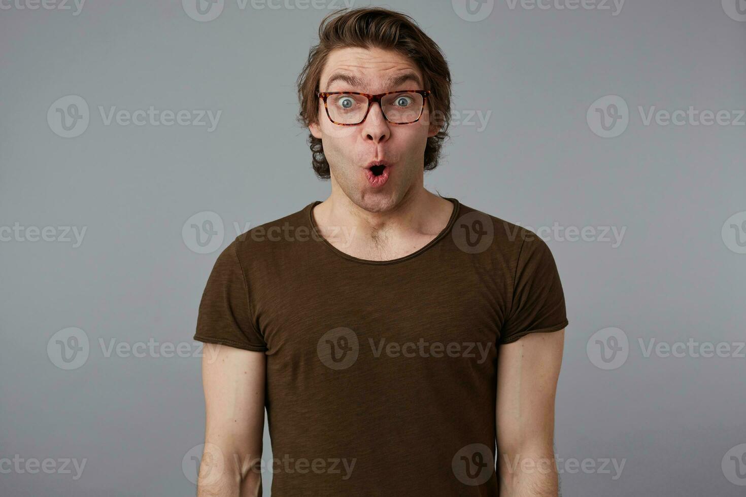 Photo of young wondered handsome man with glasses wears in basic t-shirt, stands over gray background and broadly smiles, looks happy and surprised.