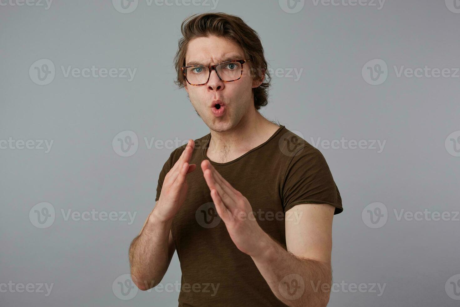 Photo of young guy with glasses wears in blank t-shirt standing in defensive posture, ready to punch, stands over gray background and try look expressing anger and fury.