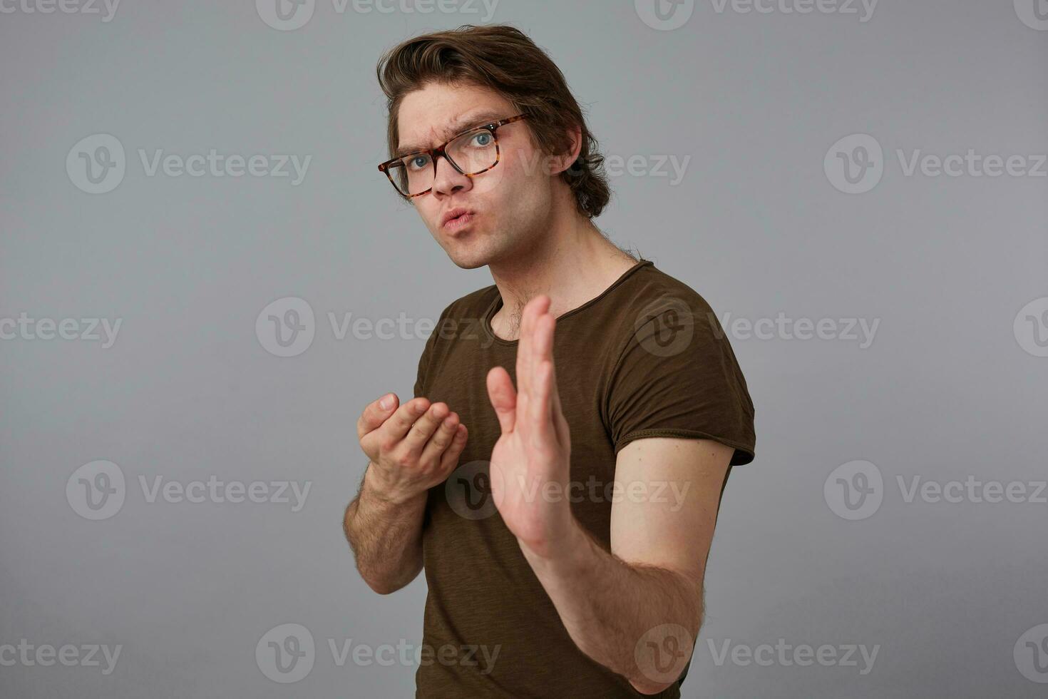 joven grave hombre con lentes usa en blanco camiseta en pie en defensivo postura, Listo a puñetazo, soportes terminado gris antecedentes y mira expresando ira y furia. foto