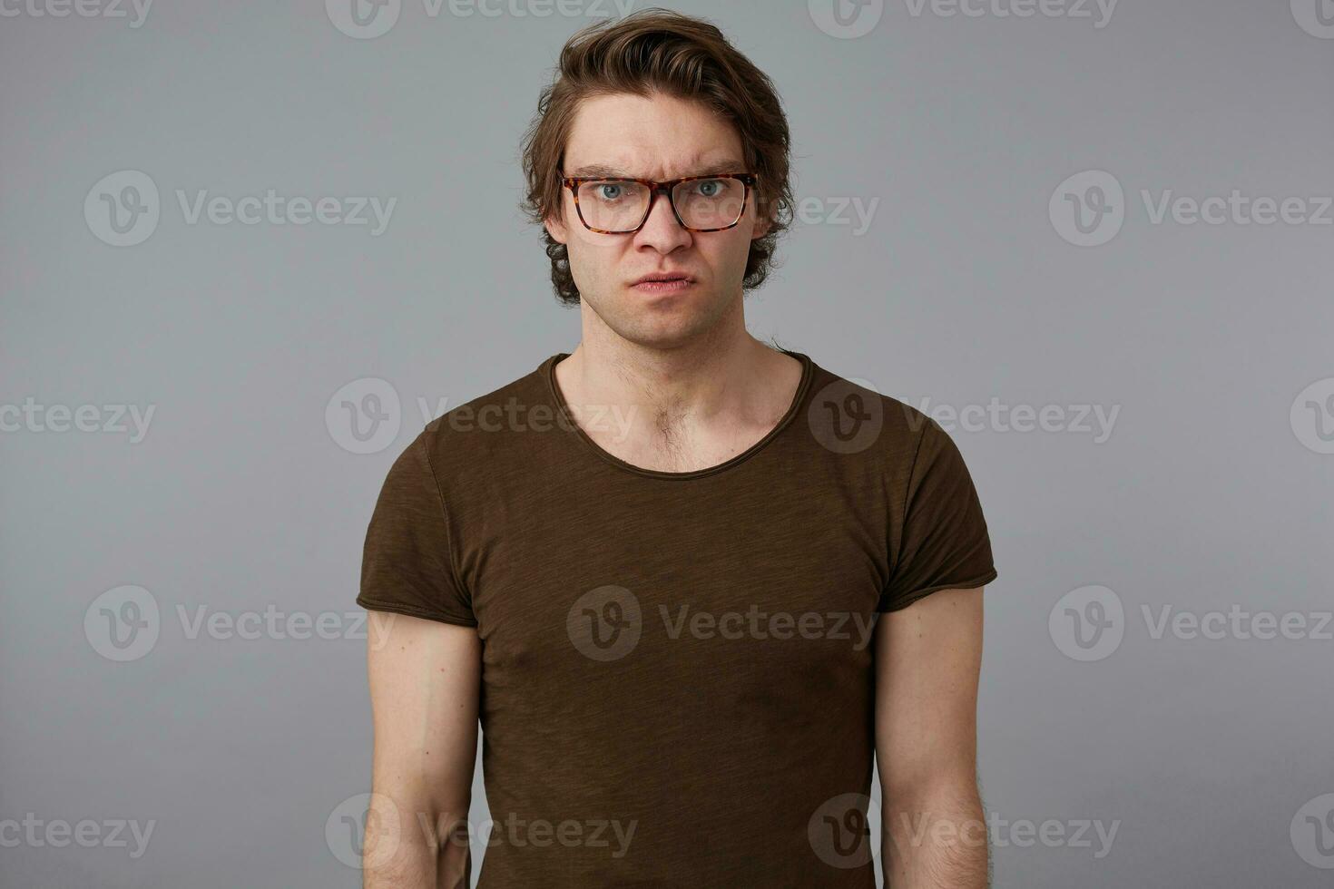 Photo of young angry guy with glasses wears in blank t-shirt, stands over gray background and evil looks at the camera.