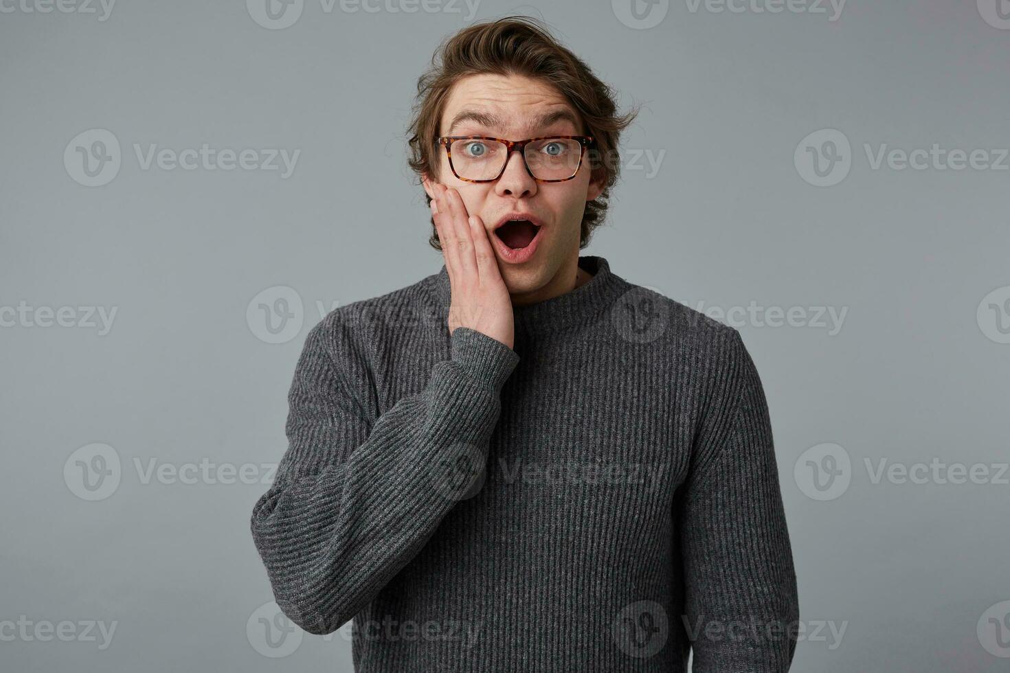 Photo of young shocked guy with glasses wears in gray sweater, stands over gray background and touches cheek, with wide open mouth and eyes, with surprised expression.