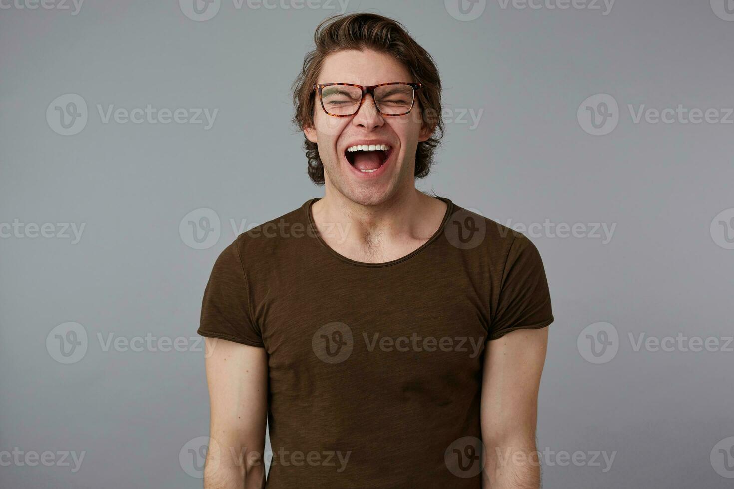 Photo of young crying guy with glasses wears in blank t-shirt, stands over gray background and looks unhappy and sad.
