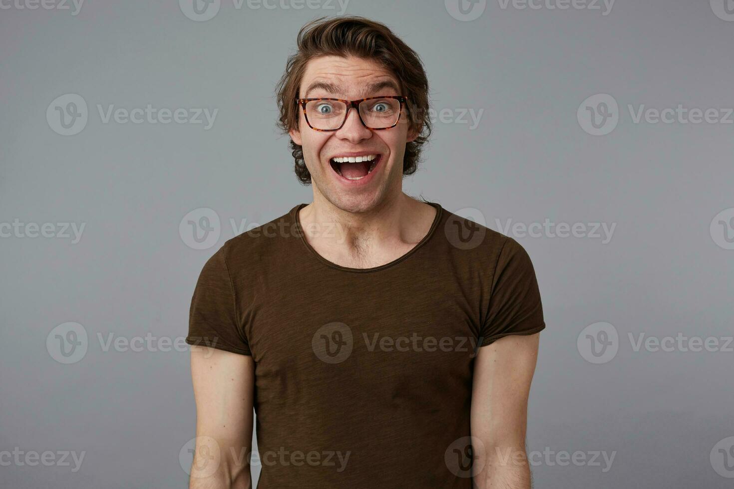 Young handsome happy amazed man with glasses wears in basic t-shirts, stands over gray background and broadly smiles, looks happy and wondered. photo