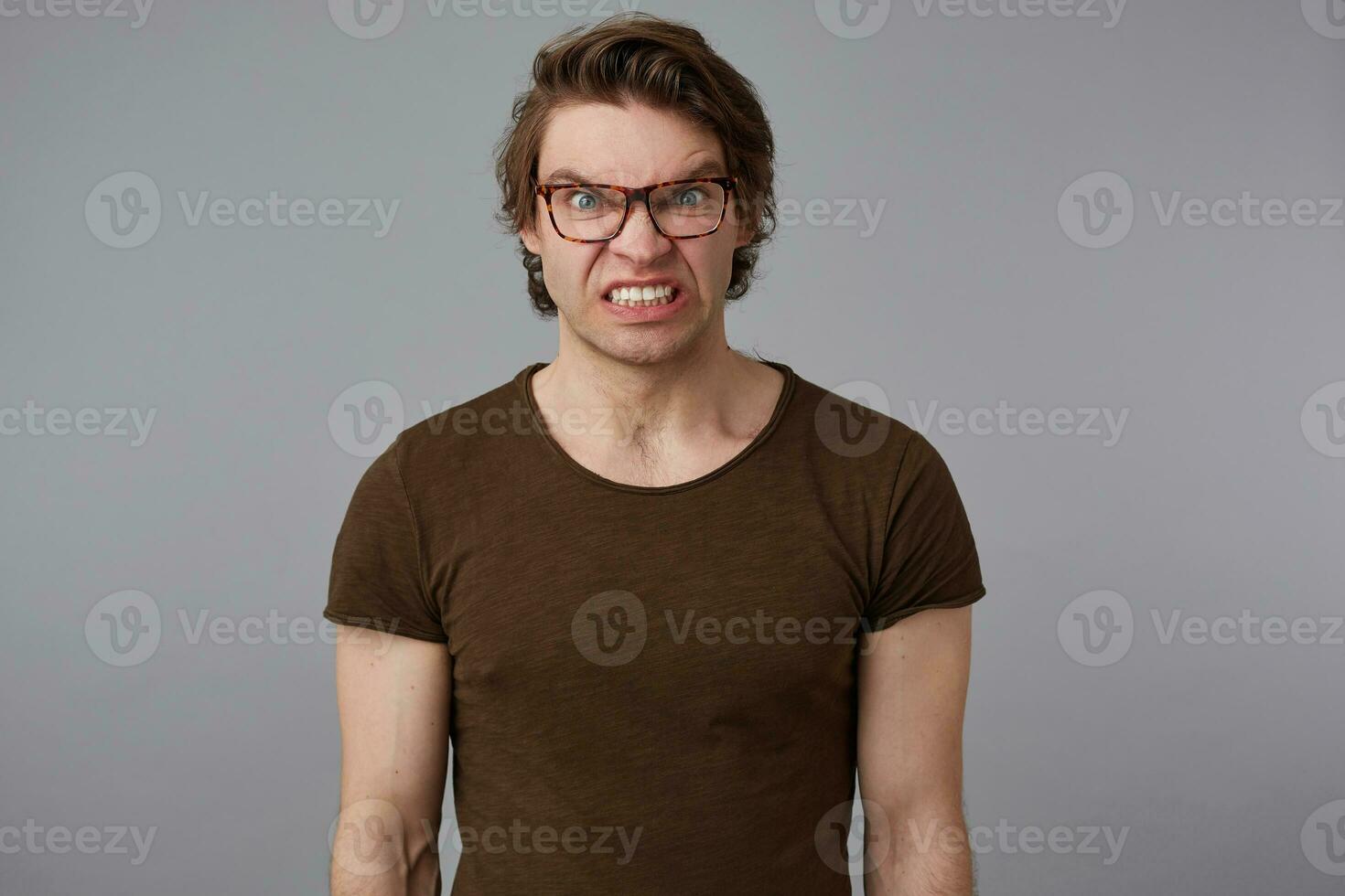 Photo of young crazy guy with glasses wears in blank t-shirt, stands over gray background and angry looks at the camera.