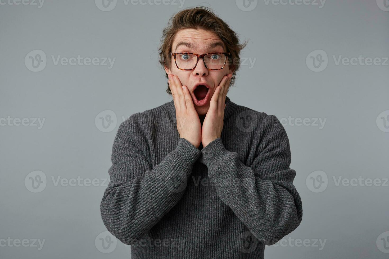 Young surprised man with glasses wears in gray sweater, stands over gray background and touches cheeks with wide open mouth and eyes, hears amazed news, with shocked expression. photo