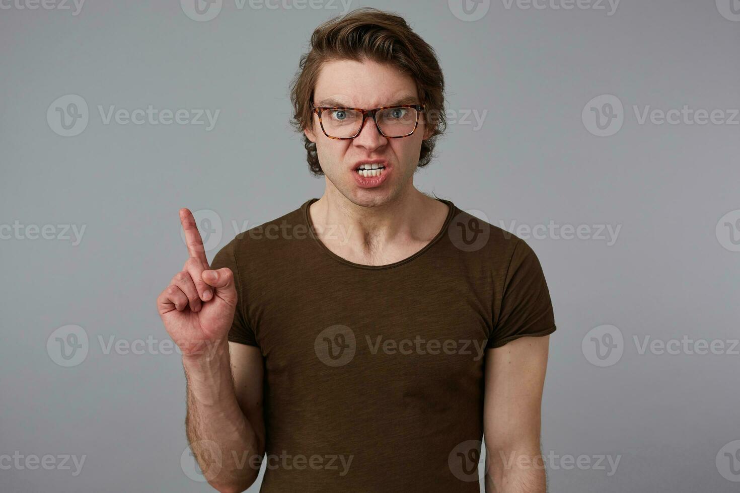 Photo of young angry guy with glasses wears in blank t-shirt, stands over gray background and looks at the camera with dissatisfied expression.