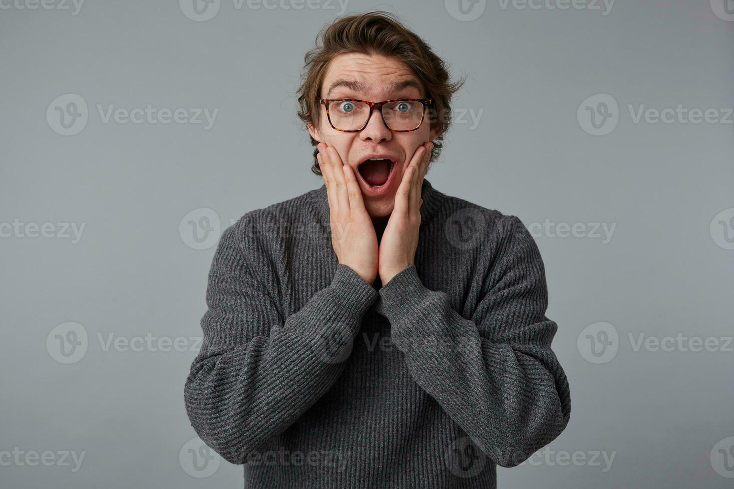 Wow, unbelievable news Young shocked guy with glasses, stands over gray background with wide open mouth and eyes, touches cheeks, with surprised expression. photo