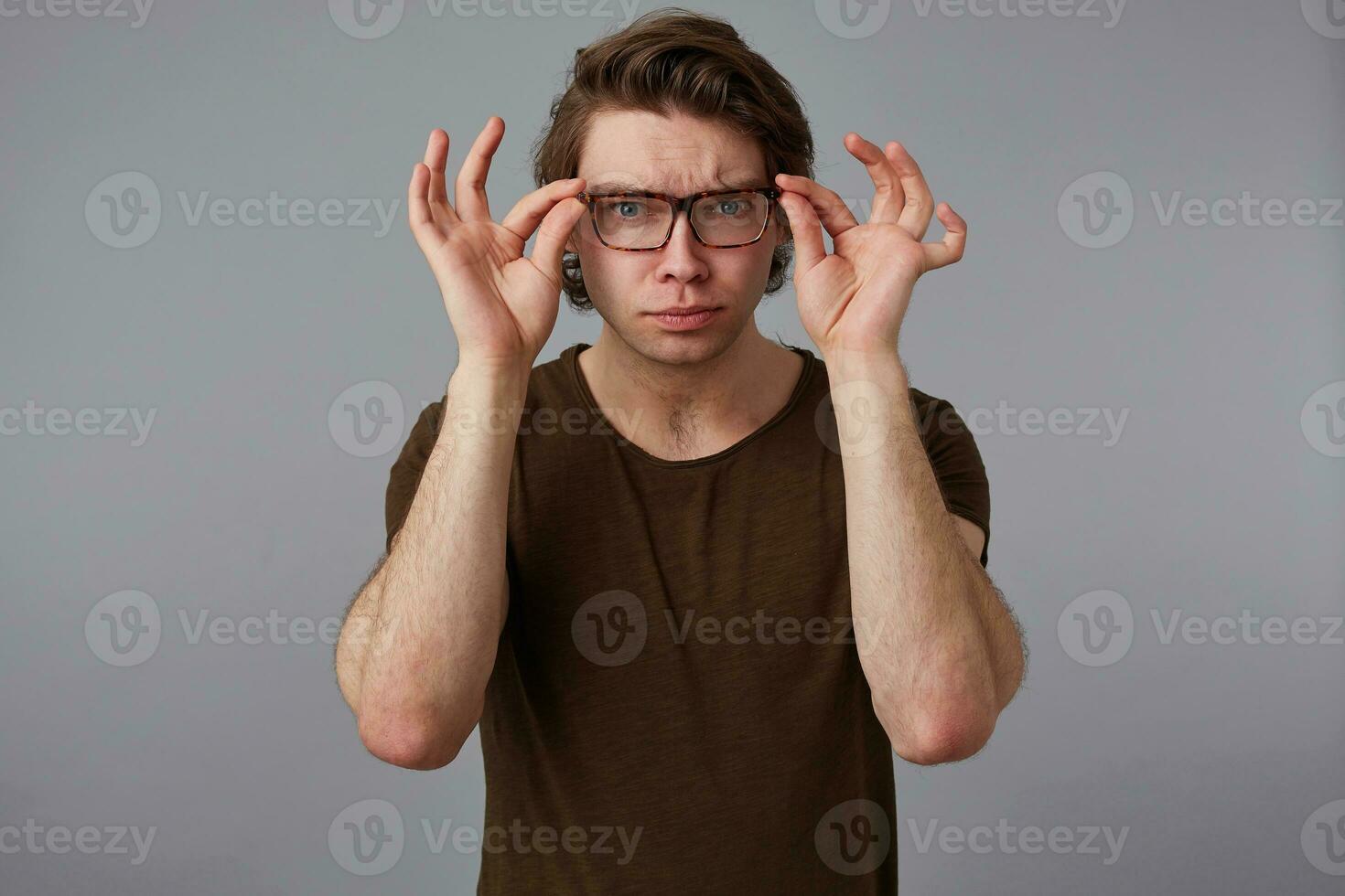 Portrait of young man with glasses wears in basic t-shirt, stands over gray background and broadly smiles, frowning looks trough glasses. photo