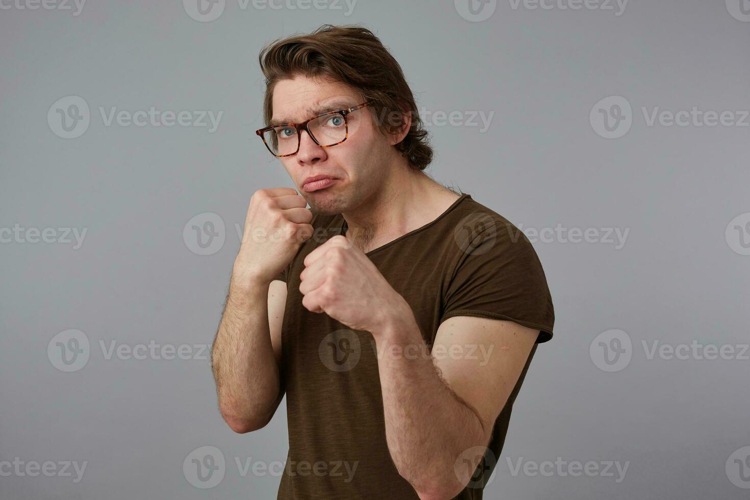 Young guy with glasses wears in blank t-shirt standing in defensive posture, keeping fists clenched, ready to punch, stands over gray background and try look expressing anger and fury. photo