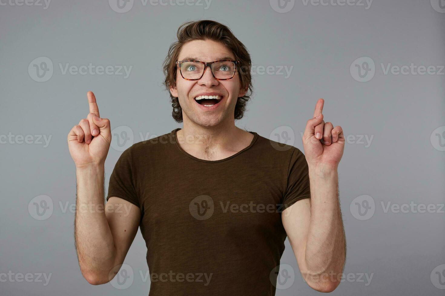 Portrait of young happy amazed man with glasses, stands over gray background with surprised expression, points fingers up at a copy space over his head. photo