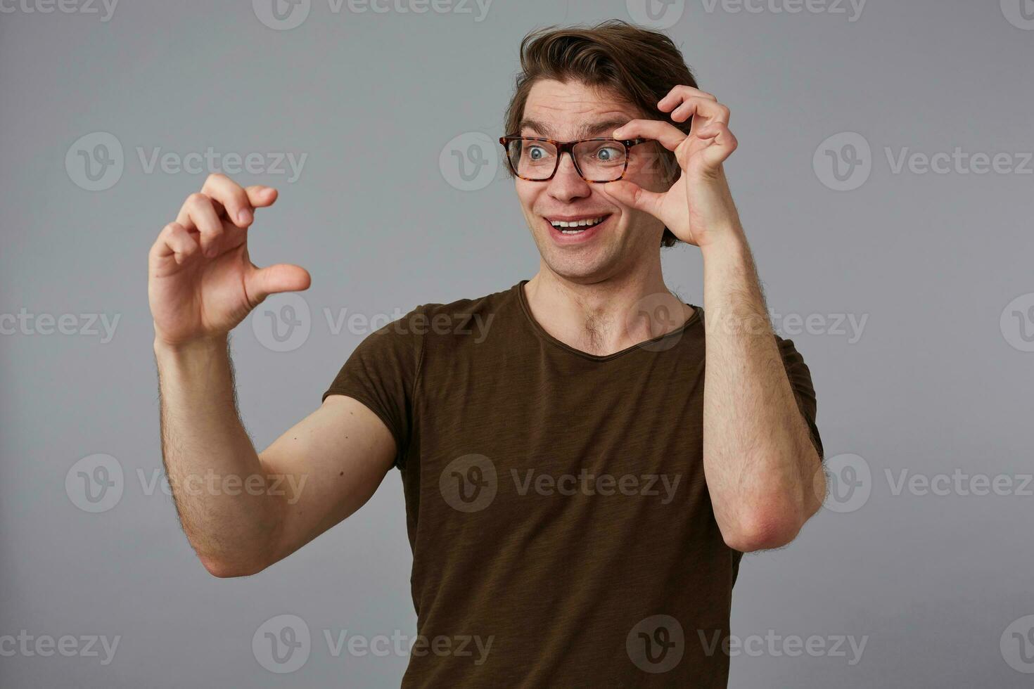 Portrait of young happy amazed man with glasses, stands over gray background looks trough glasses, shows fingers something tiny . photo