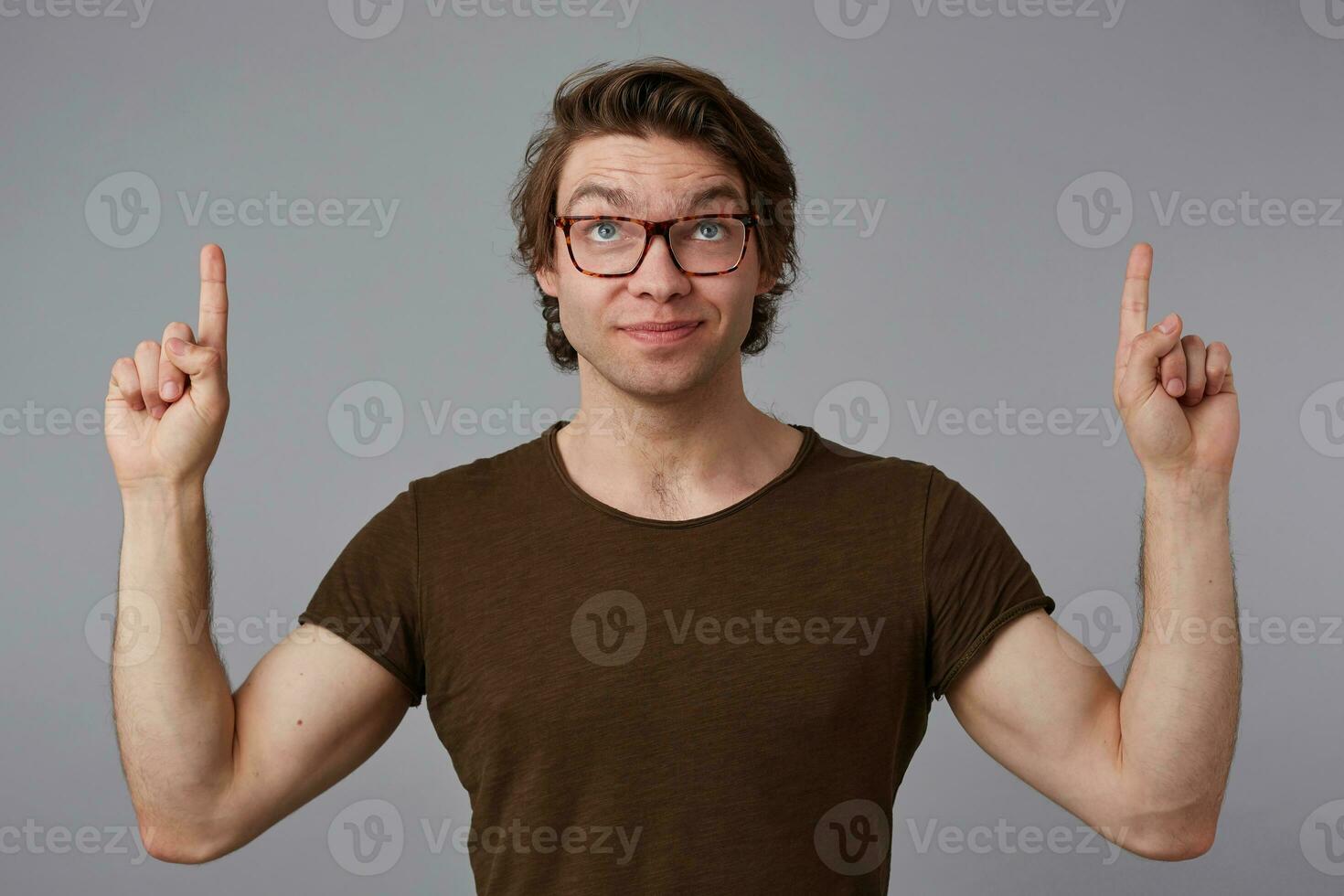 Young happy guy with glasses, stands over gray background with surprised expression, points fingers up at a copy space over his head, looks up and smiles. photo