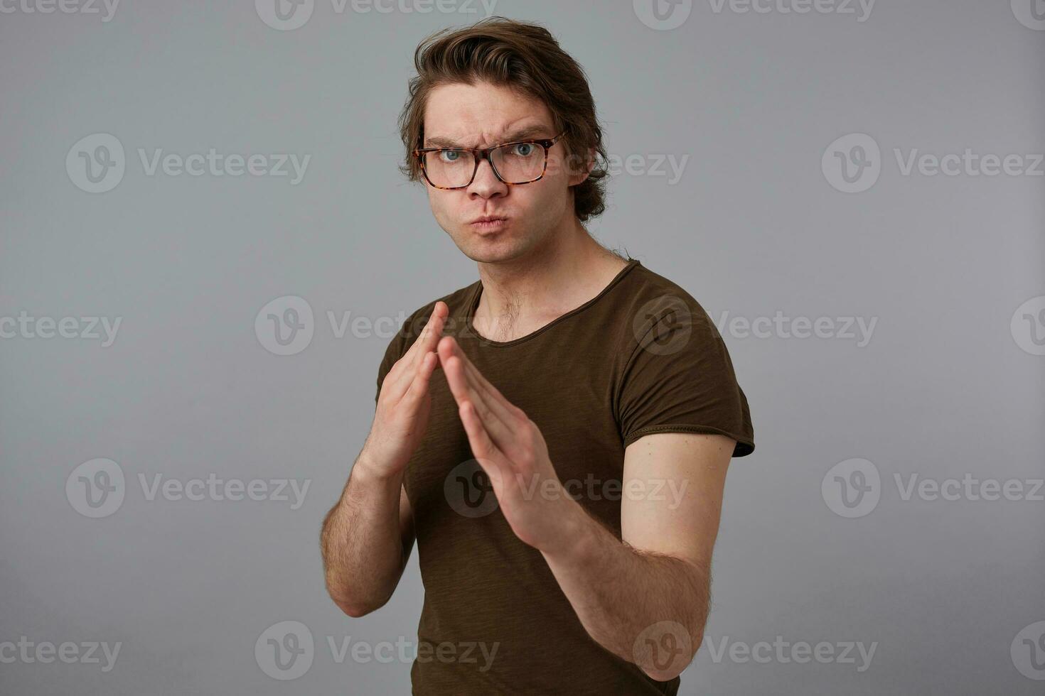 joven hombre con lentes usa en blanco camiseta en pie en defensivo postura, Listo a puñetazo, soportes terminado gris antecedentes y mira expresando ira y furia. foto