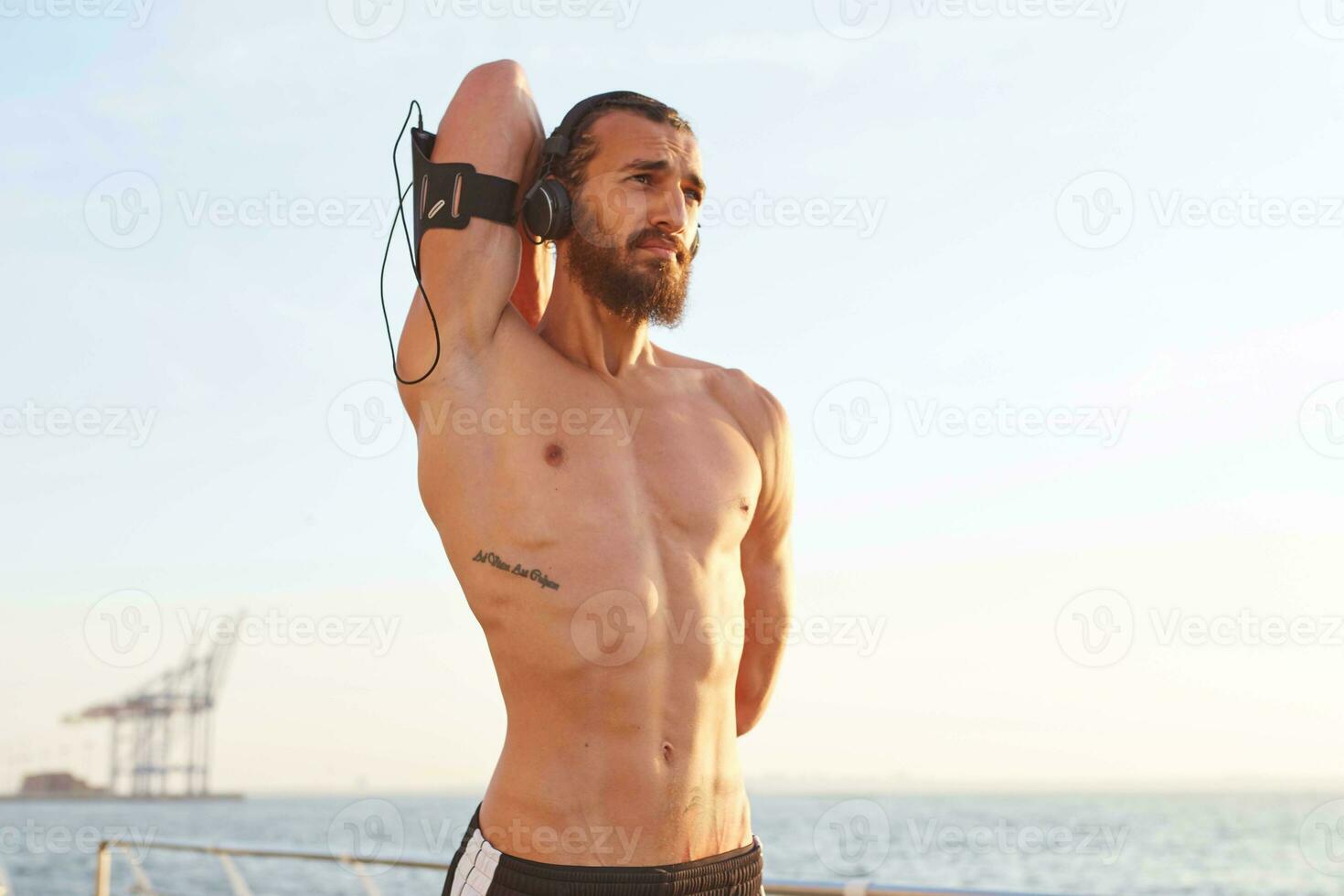 Photo of young handsome sporty man with beard. Enjoy the morning , does stretching exercises, has muscular body shape, listen favorite music on headphones.