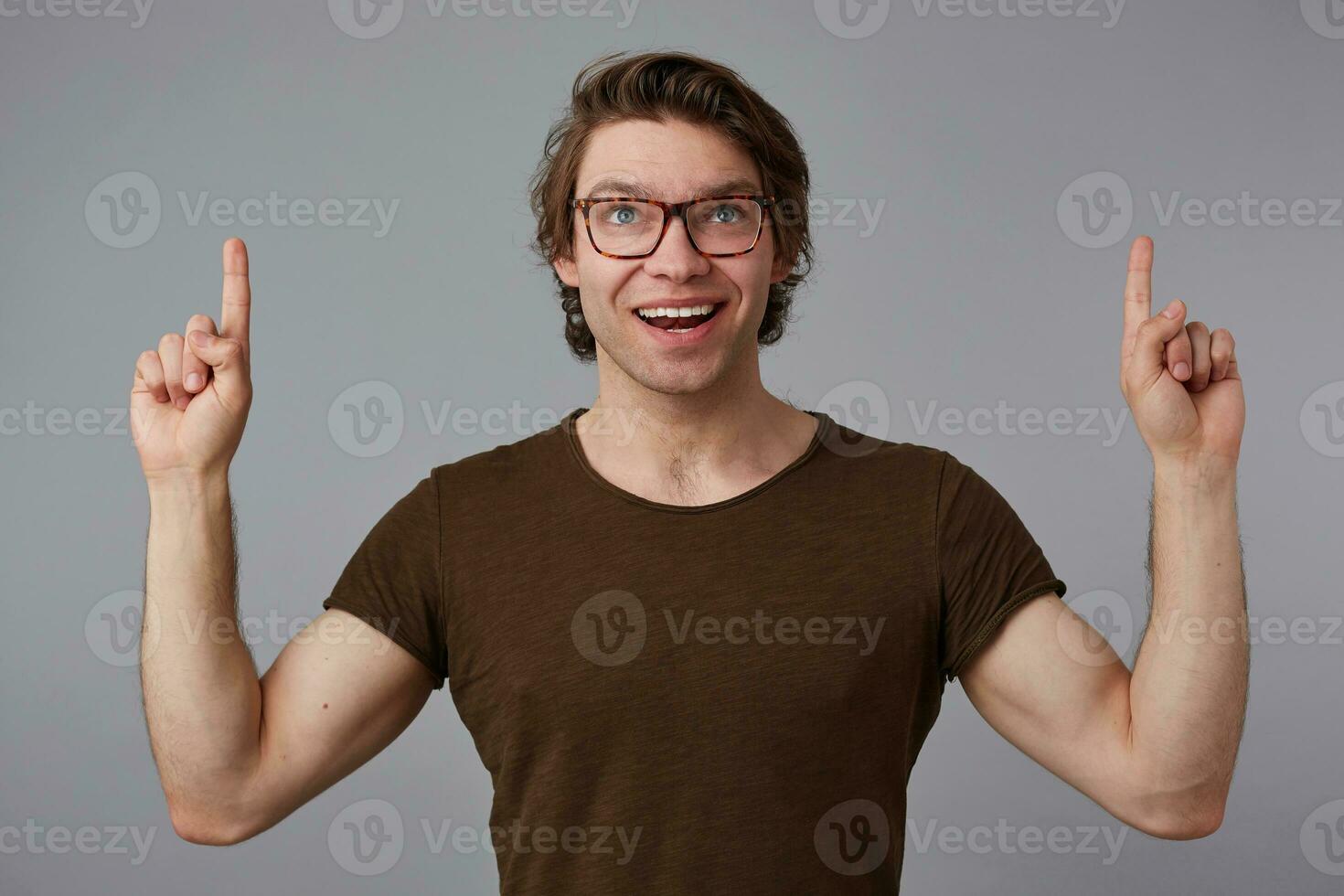 Portrait of young cheerful guy with glasses, stands over gray background with surprised expression, points fingers up at a copy space over his head, looks at the camera and broadly smiles. photo