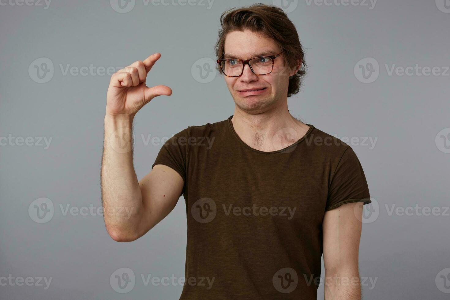 retrato de joven descontento hombre con anteojos, soportes terminado gris antecedentes mira canal anteojos, muestra dedos alguna cosa minúsculo . foto