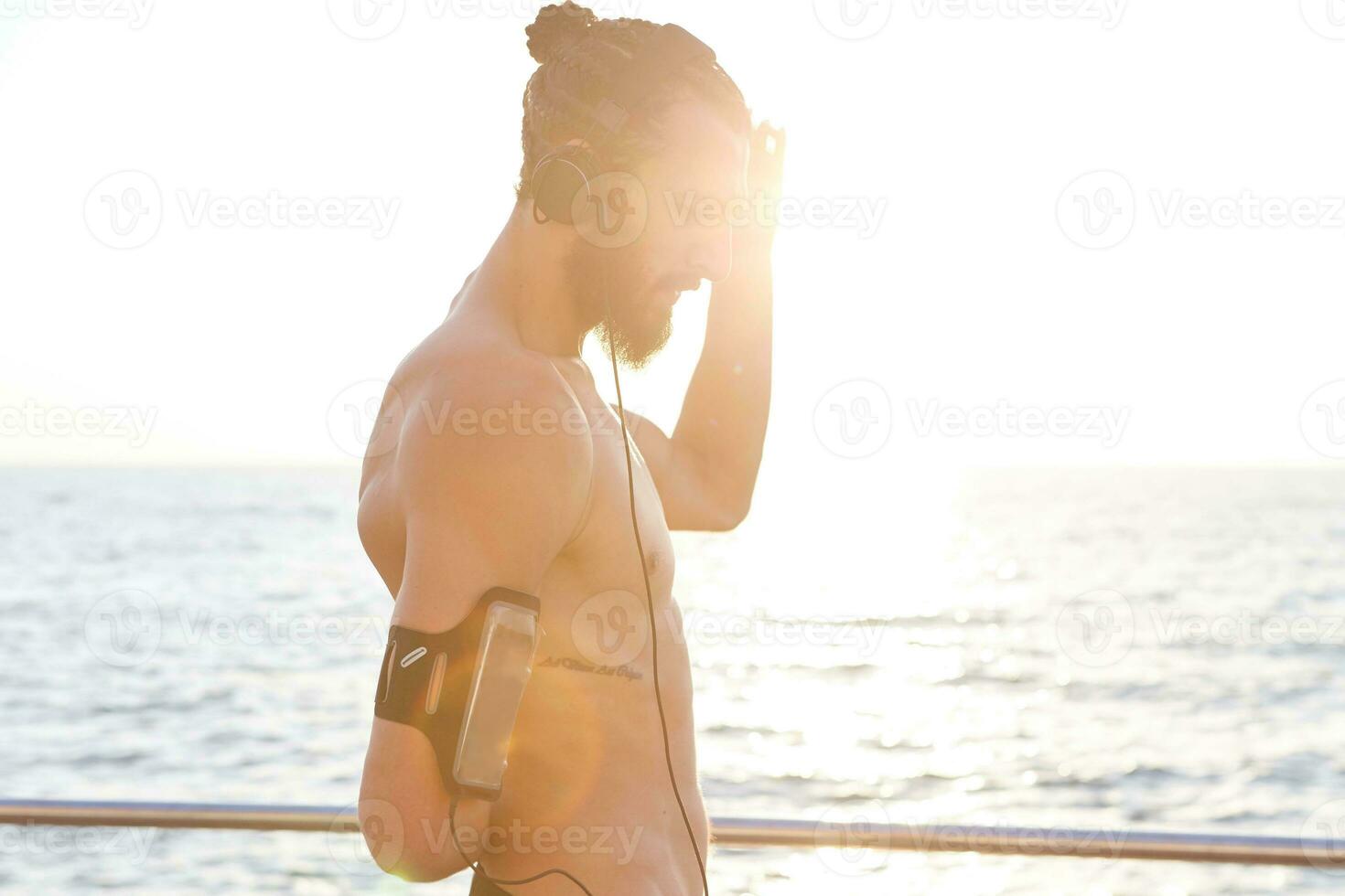 retrato de joven deportivo chico con barba, descanso después Mañana jaqueando a el playa, escucha favorito mezcla en auriculares. foto