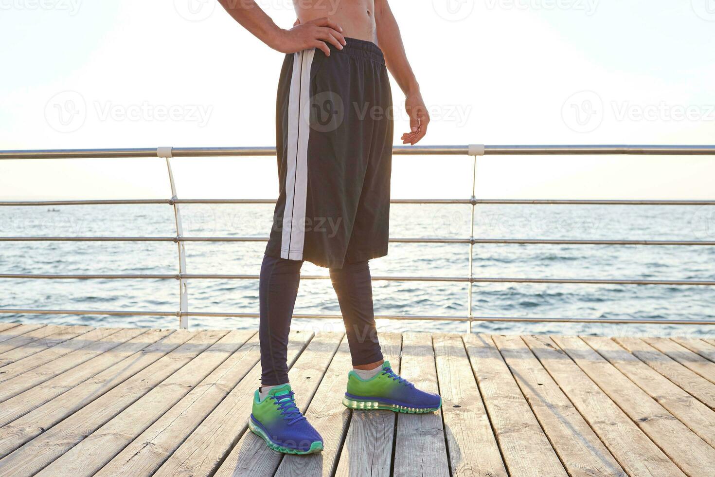 Photo of legs of sporty man, wears is colorful sneakers and striped shorts. Stand at the seaside.