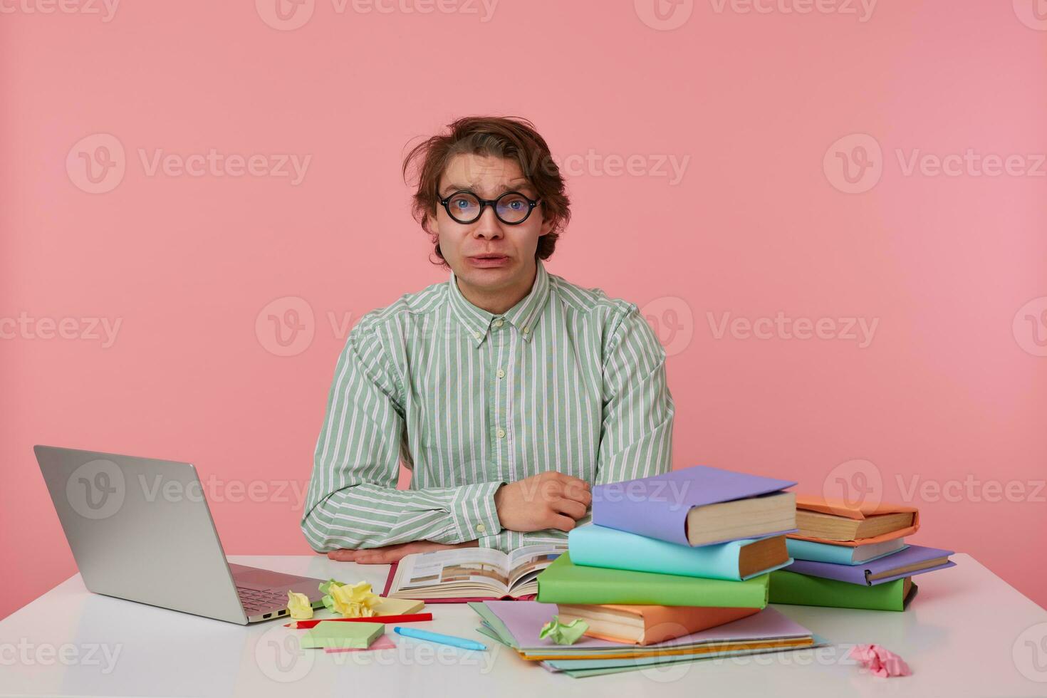 Young sad man with glasses, sitting at a table with books, working at a laptop, looks unhappy, wears on blank shirt, looks at the camera isolated over pink background. photo