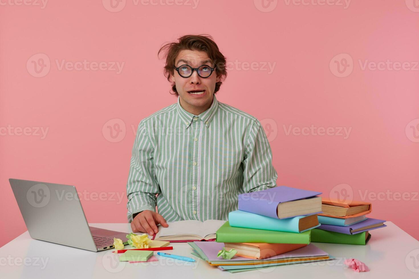 Young confused guy with glasses, wears on blank shirt, sitting at a table with books, working at a laptop, looks crazy and funny. Isolated over pink background. photo