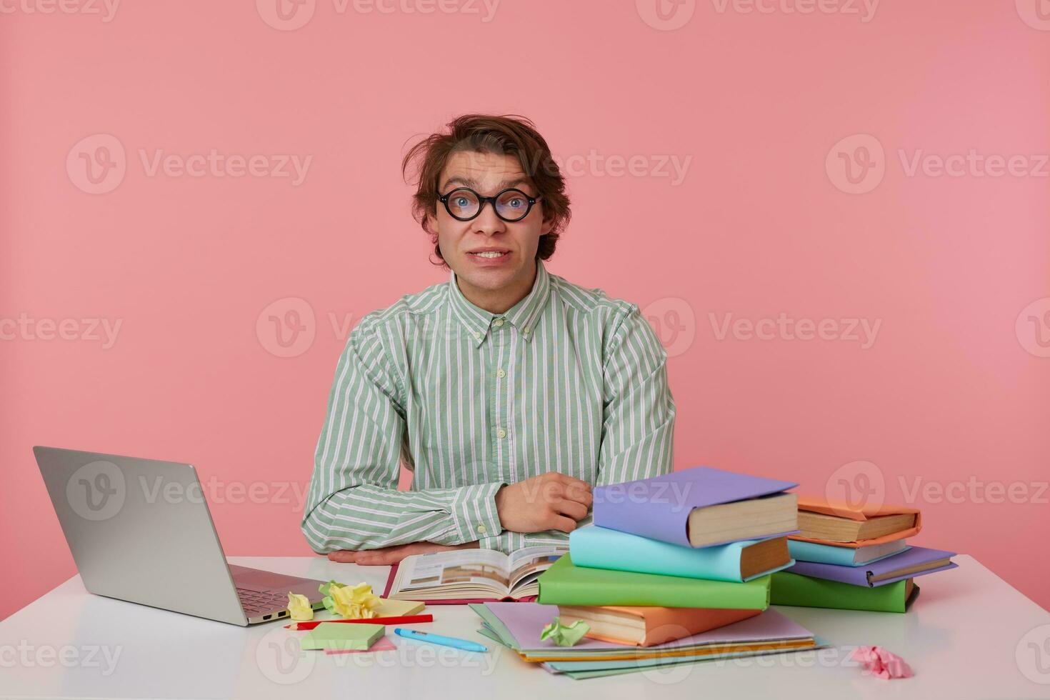 Portrait of young resentful guy with glasses, wears on blank shirt, sitting at a table with books, working at a laptop, looks confused. Isolated over pink background. photo