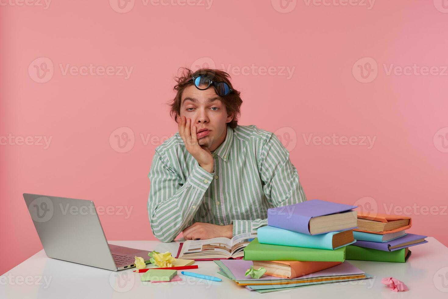 Young bored man with glasses, sitting at a table with books, working at a laptop, looks sleepy, wears on blank shirt, tiredly looks at the camera isolated over pink background. photo
