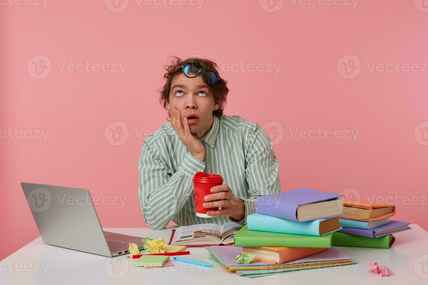 Portrait of young bored man with glasses, sitting at a table with books, working at a laptop, looks tired, wears on blank shirt, looks up isolated over pink background. photo