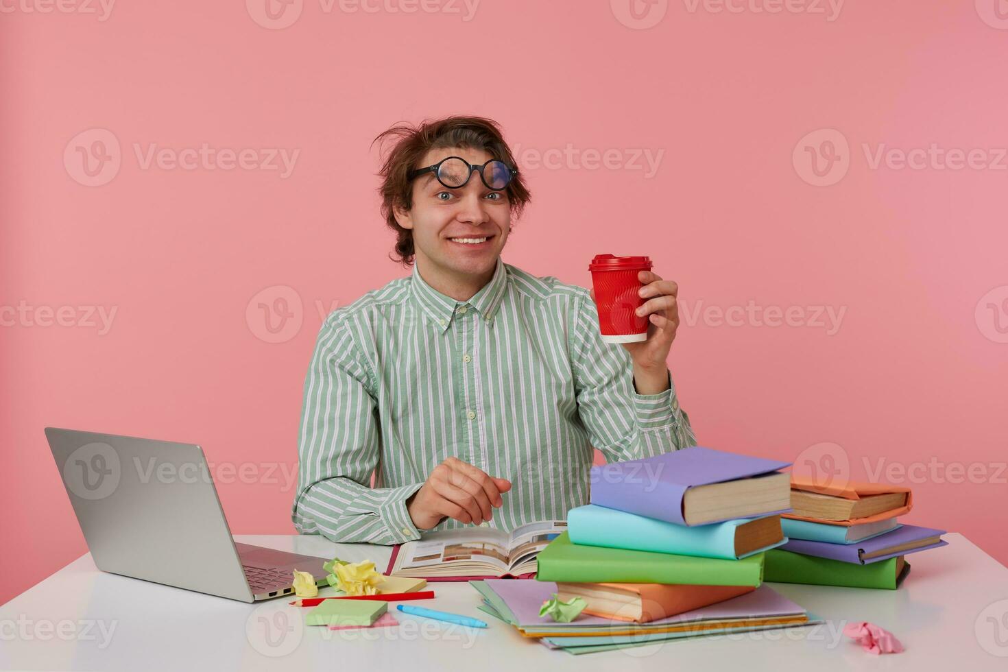 young happy man with glasses, sitting at a table with books, working at a laptop, looks glad, wears on blank shirt, looks at the camera and holds cup of coffe isolated over pink background. photo