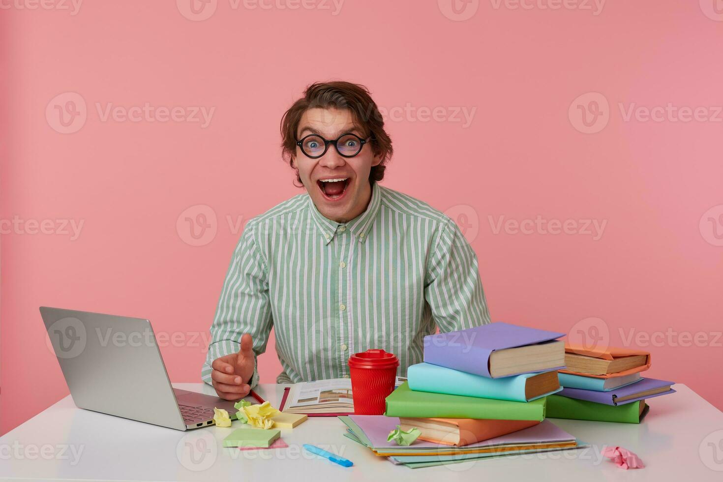 young cheerful man with glasses, sitting at a table with books, working at a laptop, looks glad, wears on blank shirt, looks at the camera with happy expression, isolated over pink background. photo