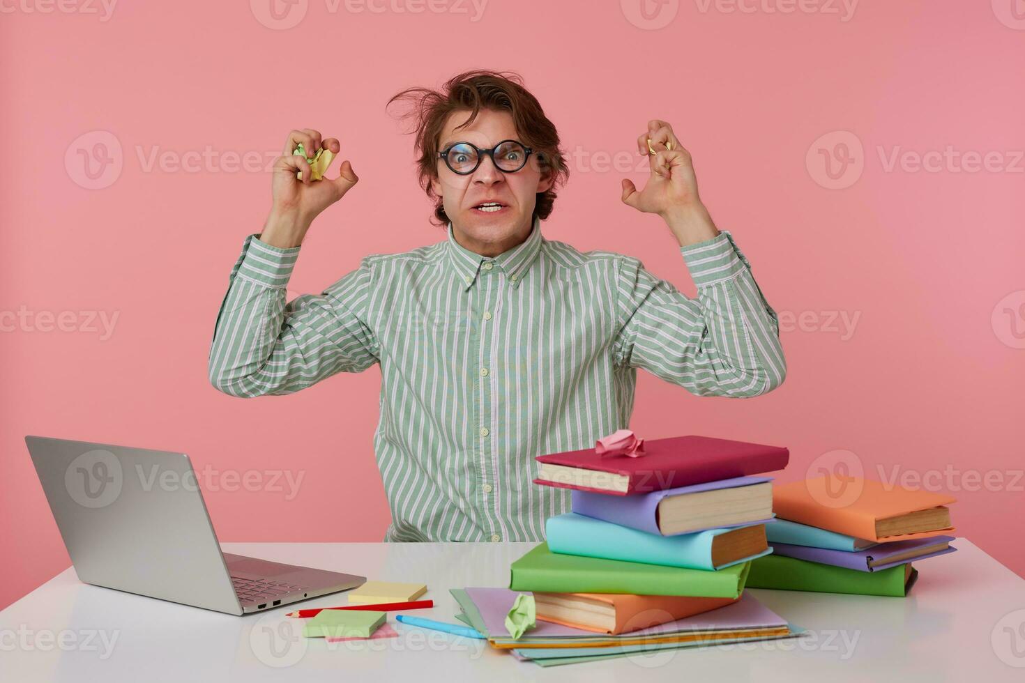 Young angry guy with glasses, wears on blank shirt, sitting at a table with books, working at a laptop, looks crazy and dissatisfied, something wrong. Isolated over pink background. photo