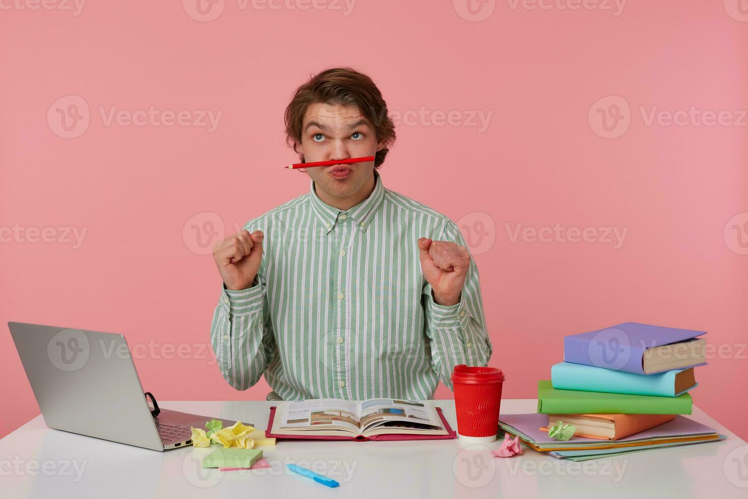 Photo of young funny guy with glasses, sitting at a table with books, working at a laptop, looks up and holds pencil at the lips, with up fists, isolated over pink background.