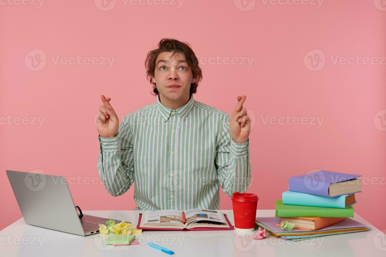 Portrait of young guy with glasses, sitting at a table with books, working at a laptop, looks up and wishes, with crossed fingers hopes for good luck, isolated over pink background. photo