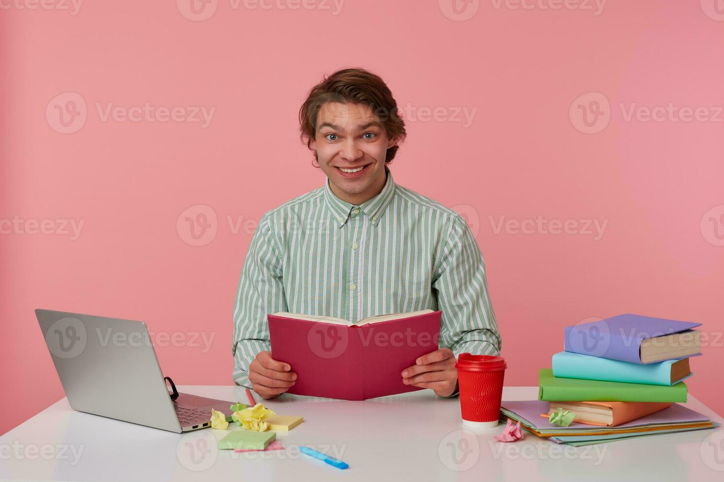 Photo of young cheerful guy with glasses, sitting at a table with books, working at a laptop, holding open book, looks at the camera and smiling, isolated over pink background.