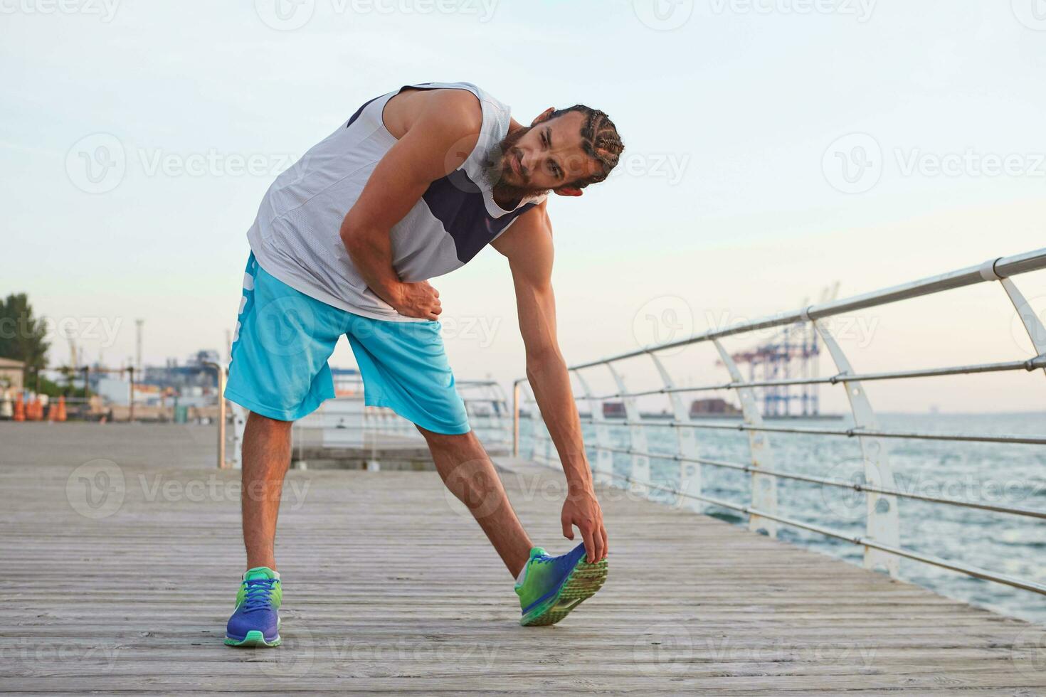 Photo of young sporty bearded guy doing stretching, morning exercises by the sea, warm-up after run, leads healthy active lifestyle. Fitness and healthy concept.