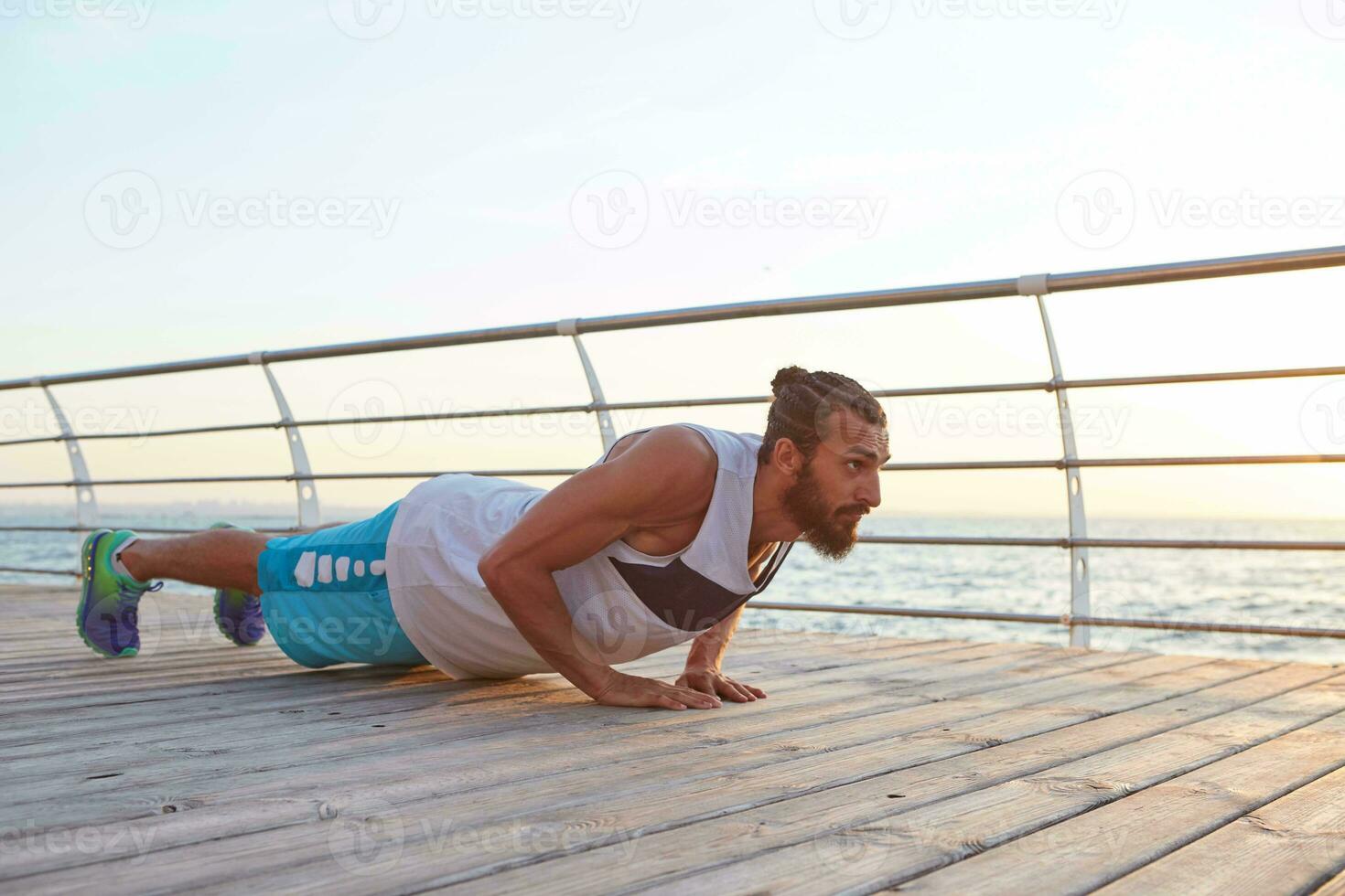 Young bearded athletic gay doing pushups, keeps the plank, doing morning exercises by the sea, warm-up after run, leads healthy active lifestyle. Fitness and healthy concept. photo