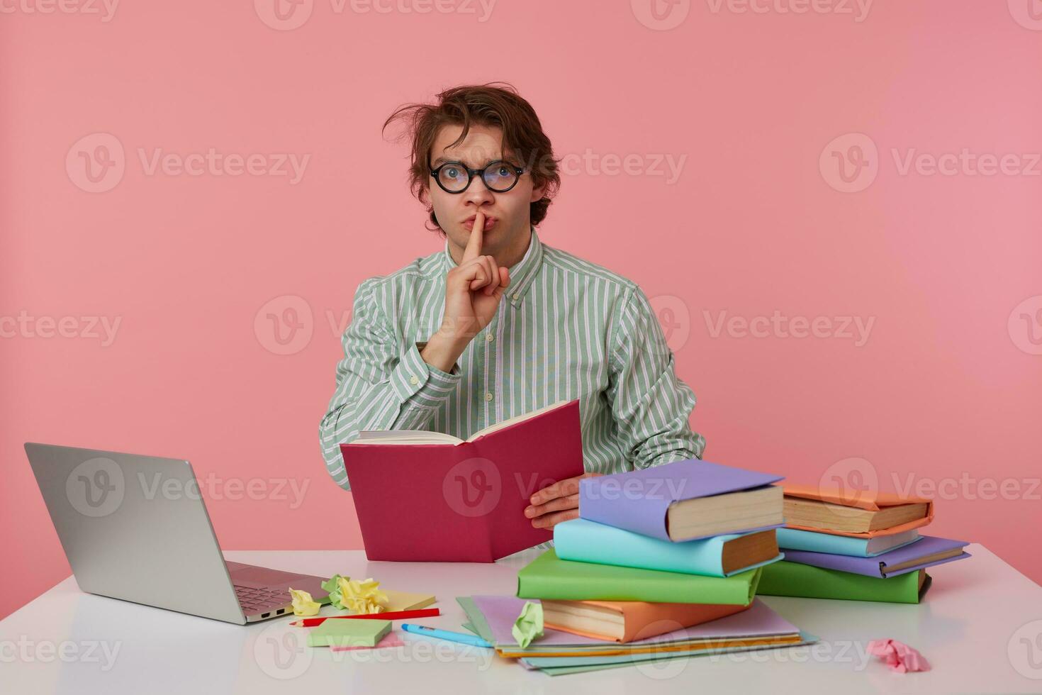 man guy with glasses, wears on blank shirt, sitting at a table with books and laptop laptop, holds a book in his hands and asks to keep quiet, shows silence gesture. Isolated over pink background. photo