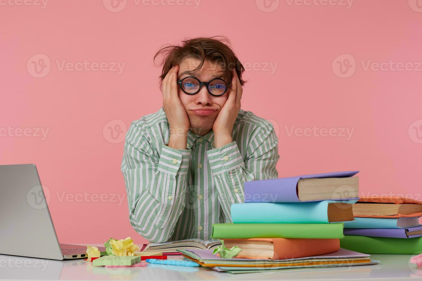 joven cansado hombre con gafas,sentado a un mesa con libros, trabajando a un computadora portátil, mira infeliz y triste, tristemente mira arriba y Sueños de dejando hogar como pronto como posible, aislado terminado rosado antecedentes. foto