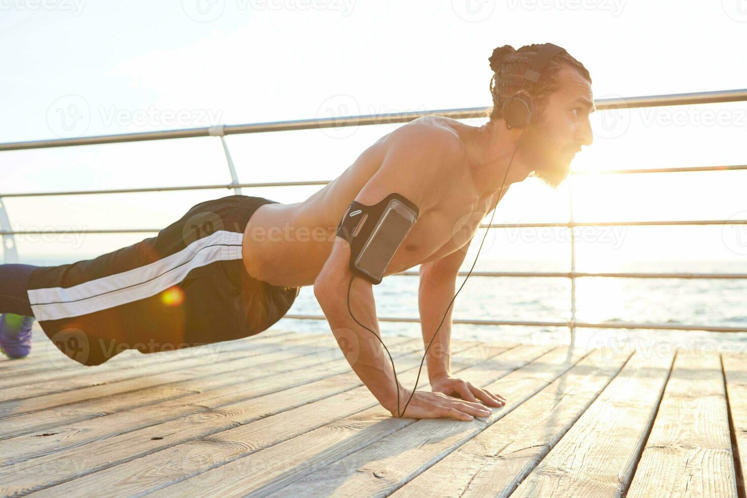 Photo of young bearded man doing morning exercises by the sea, listening favorite songs on headphones, doing pushups, keeps the plank, warm-up after run, leads healthy active lifestyle.