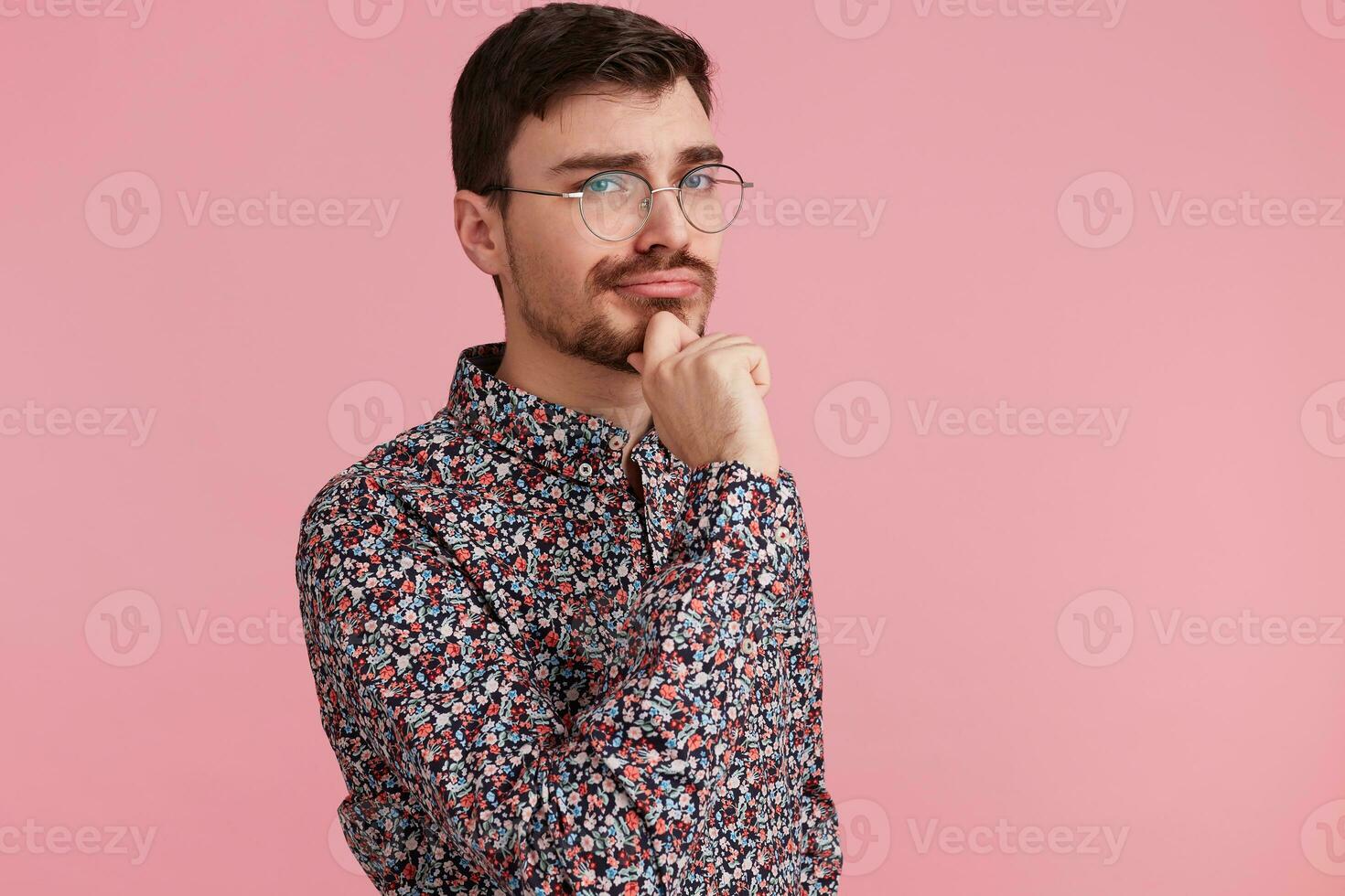 Portrait of young man in colorful shirt looking upwards, copy space on the right side, think about problem, while touches cheek, isolated over pink background. photo